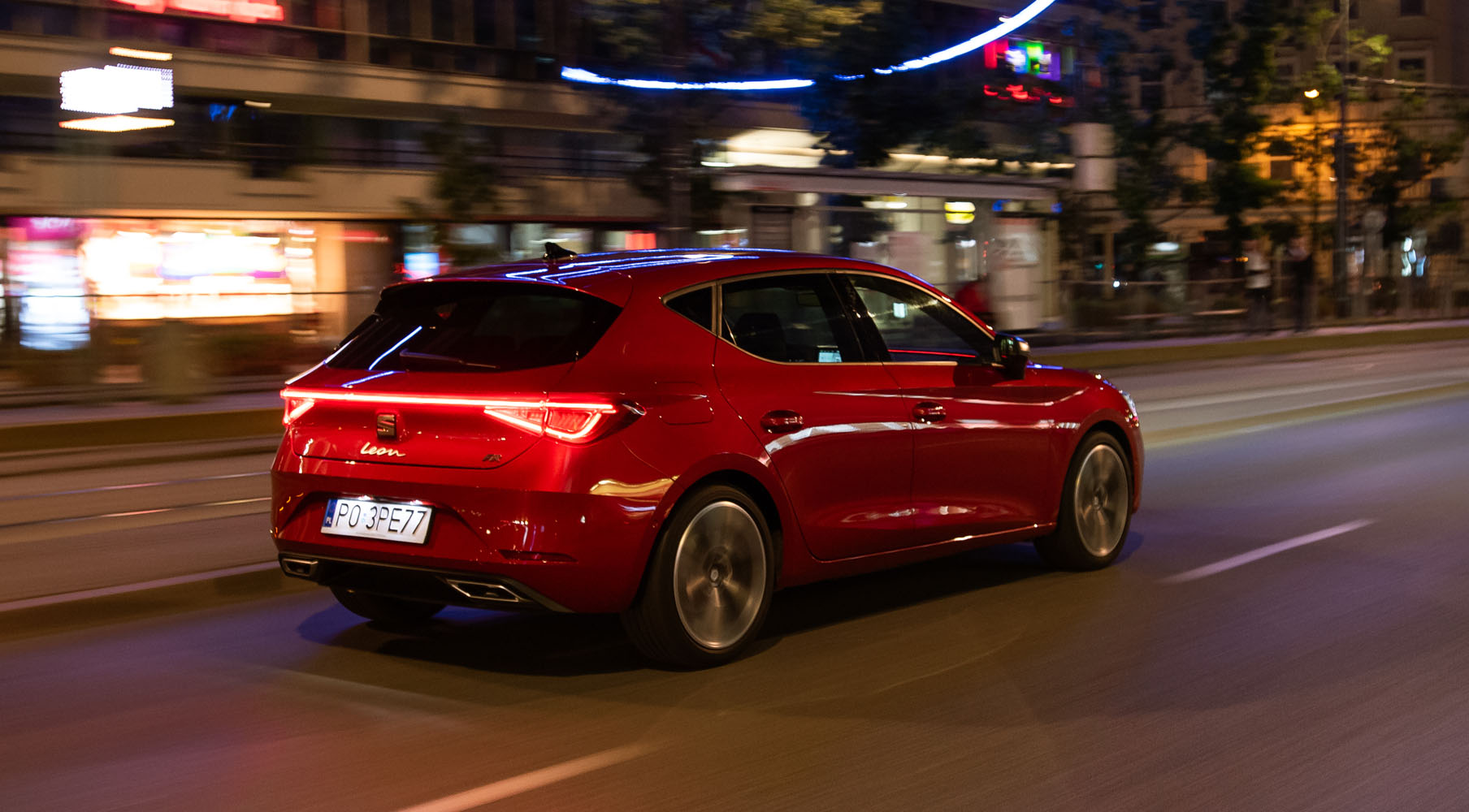 Seat Leon w kolorze Desire Red  przejeżdżający przez nocne miasto z zapalonymi tylnymi światłami LED.