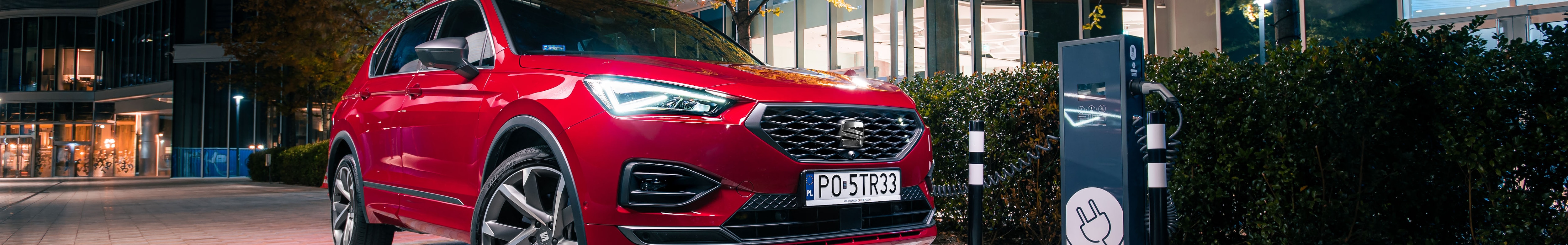 Elektryczny Seat Tarraco w kolorze Desire Red z zapalonymi przednimi światłami LED podłączony do miejskiej stacji ładowania.