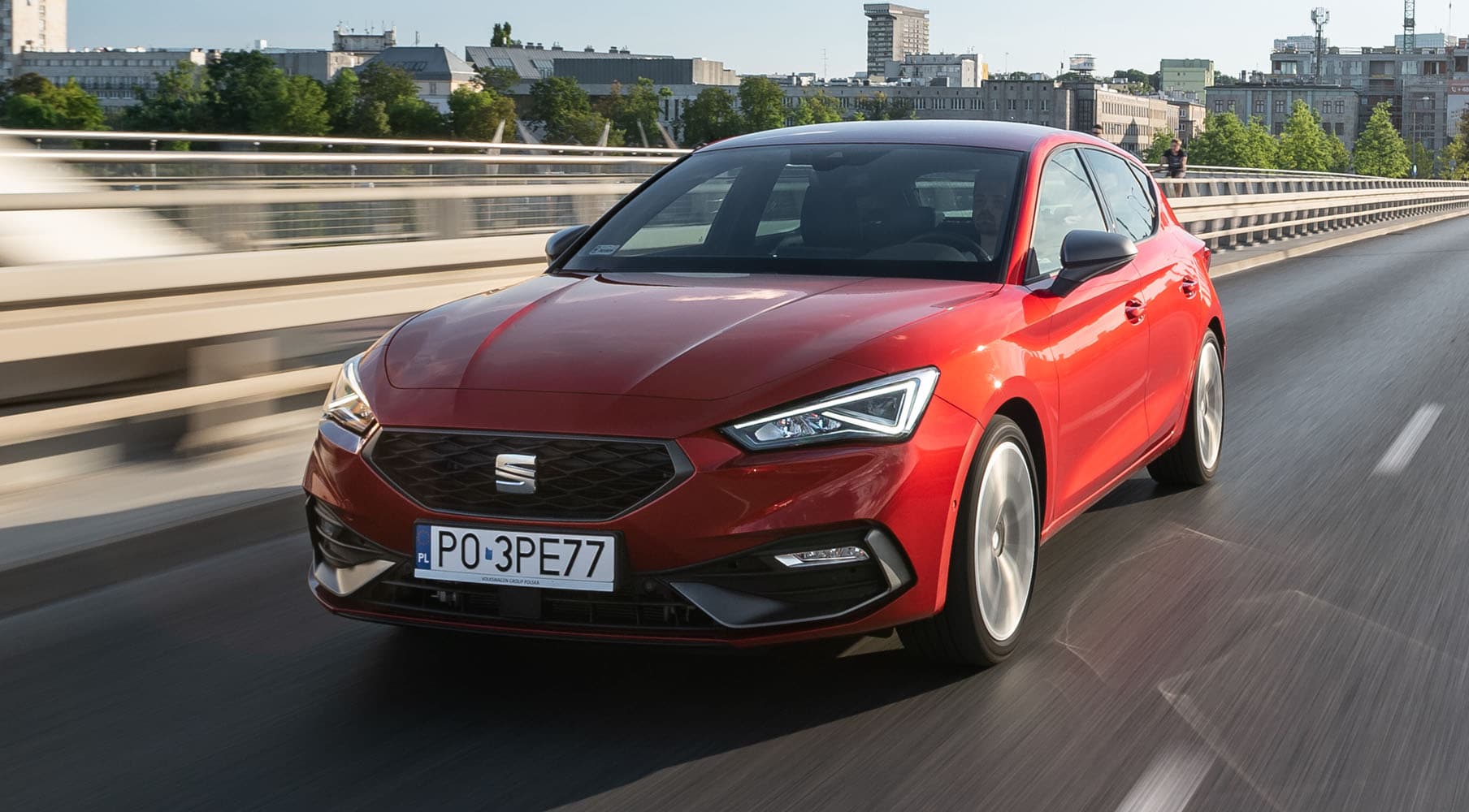 Seat Leon w kolorze Desire Red  przejeżdżający przez most podczas słonecznego dnia w mieście. 