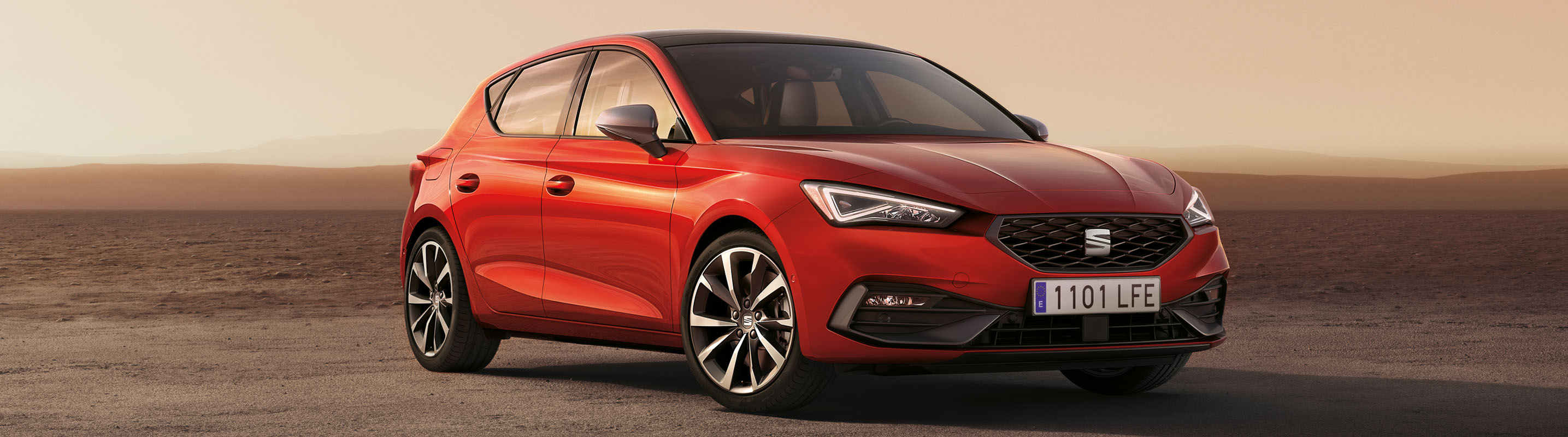 Zaparkowany Seat Leon w kolorze Desire Red z zapalonymi przednimi światłami LED, na piaszczystej drodze podczas zachodu słońca. 