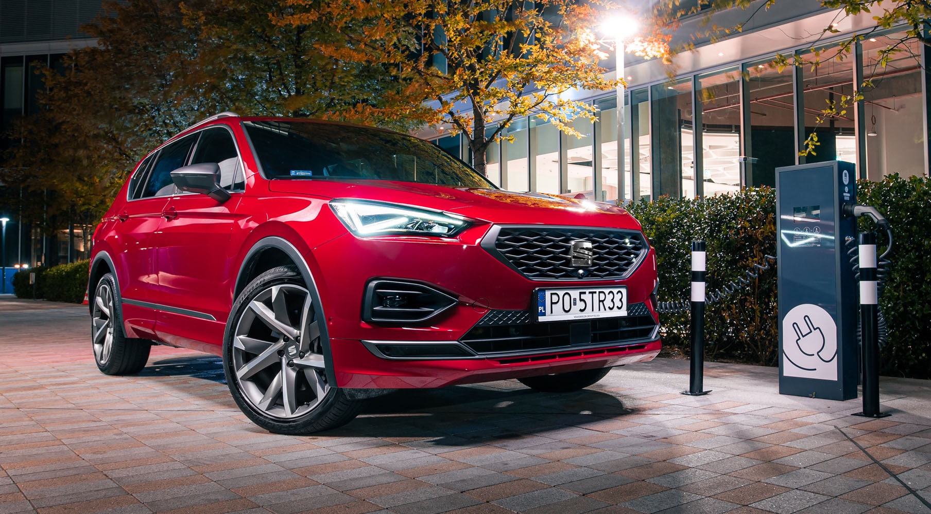 Elektryczny Seat Tarraco w kolorze Desire Red z zapalonymi przednimi światłami LED podłączony do miejskiej stacji ładowania.