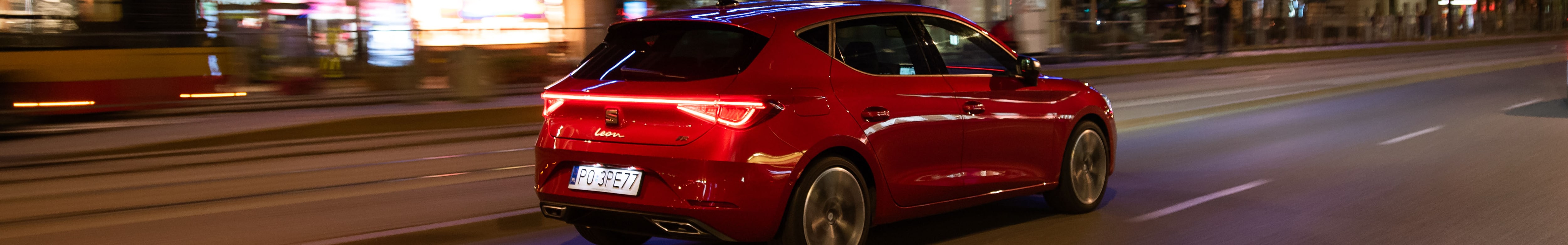 Seat Leon w kolorze Desire Red  przejeżdżający przez nocne miasto z zapalonymi tylnymi światłami LED.