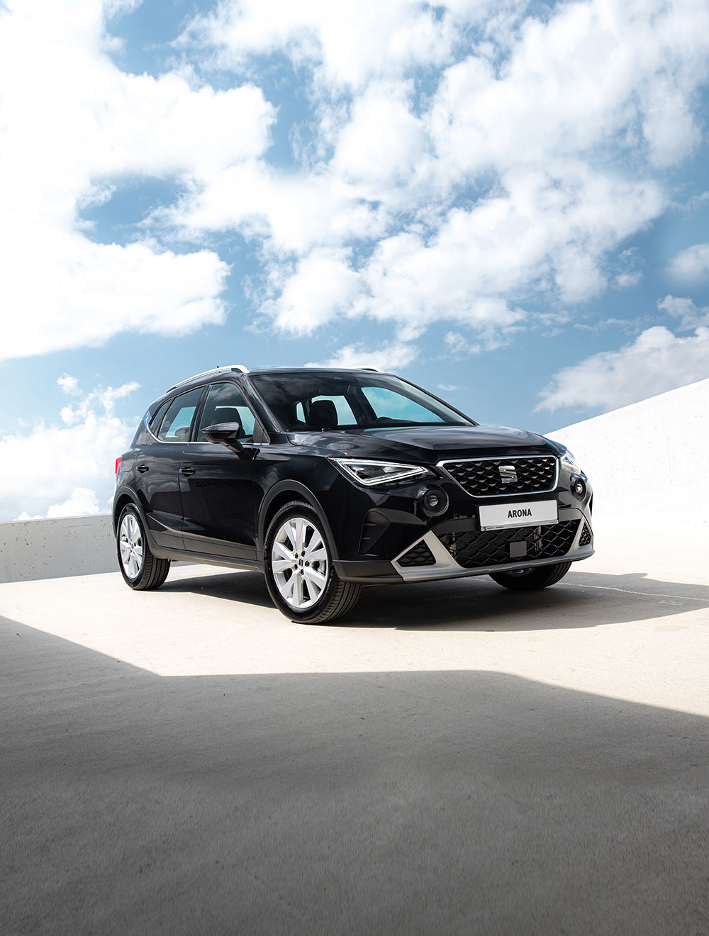 Kobieta idzie z rośliną w stronę SEAT-a Arona w kolorze dark camouflage z dachem w kolorze candy white