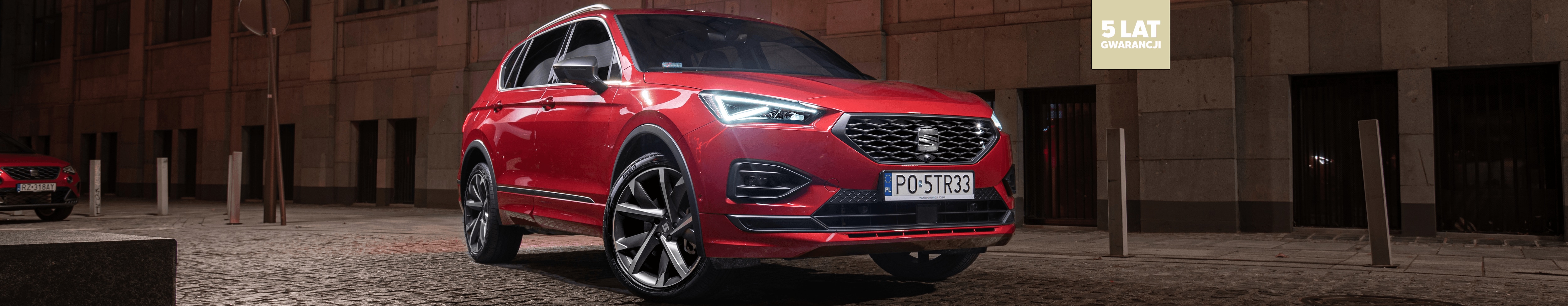 SEAT Tarraco w kolorze Desire Red, metalizowany. Auto w ruchu, zapalone reflektory LED. Widok na lewą stronę z przodu.