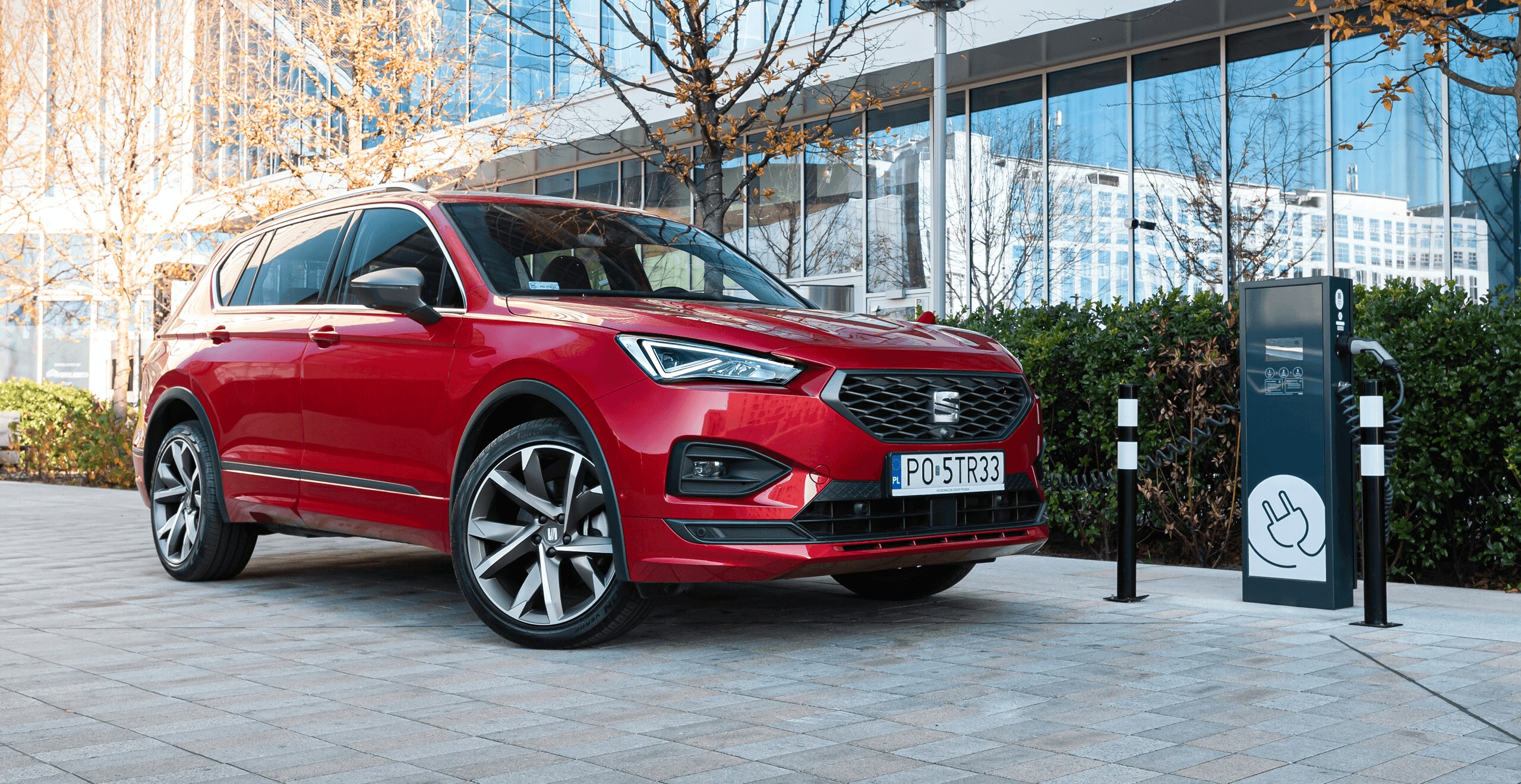 Hybrydowy SEAT Tarraco w kolorze Desire Red, metalizowany ze stacją ładowania samochodów w tle