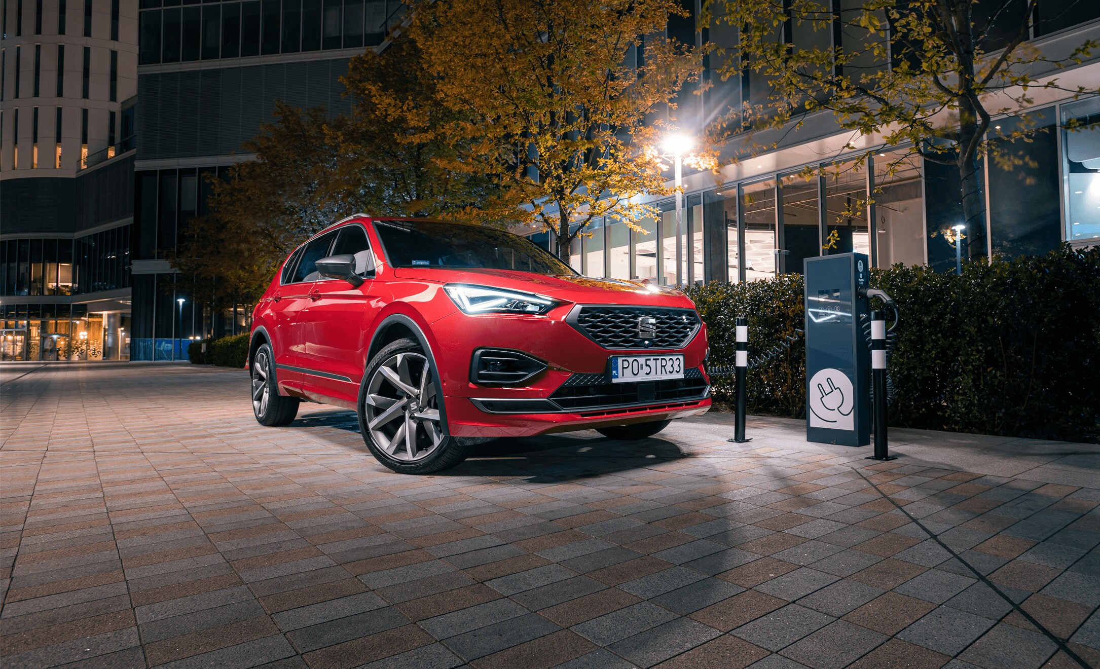 Hybrydowy SEAT Tarraco w kolorze Desire Red,  ze stacją ładowania samochodów w tle. Miasto nocą. 