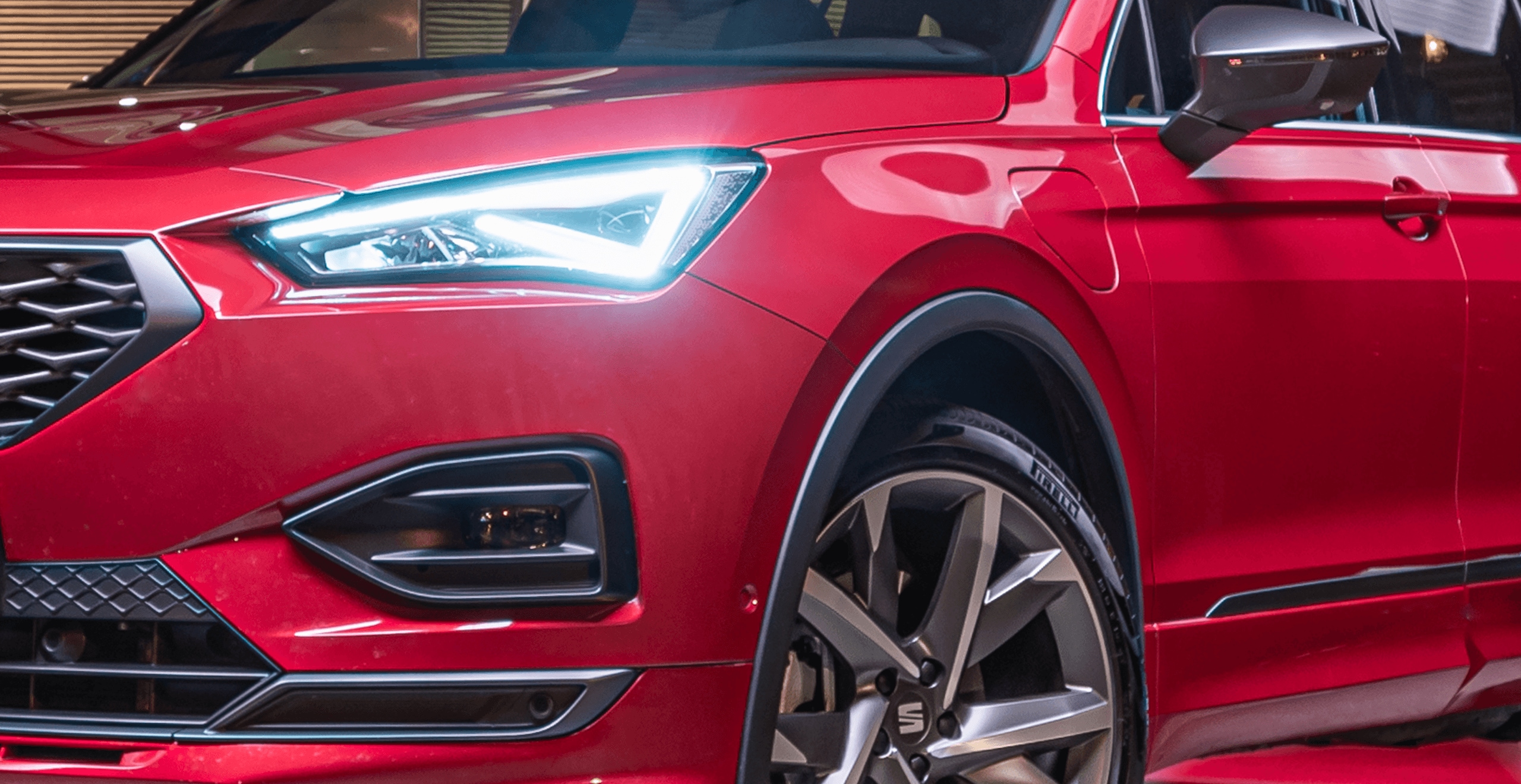 SEAT Tarraco w kolorze Desire Red, metalizowany. Widok na lewą stronę z przodu. Na pierszym planie zapalone przednie światło – reflektor LED. Auto w ruchu, miasto nocą.