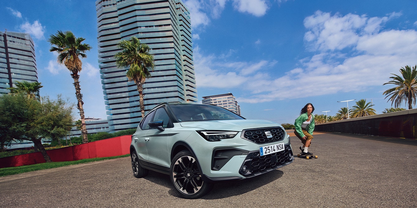 SEAT Arona zaparkowany na słonecznej ulicy na tle nowoczesnej miejskiej zabudowy, obok której przejeżdża deskorolkarz.
