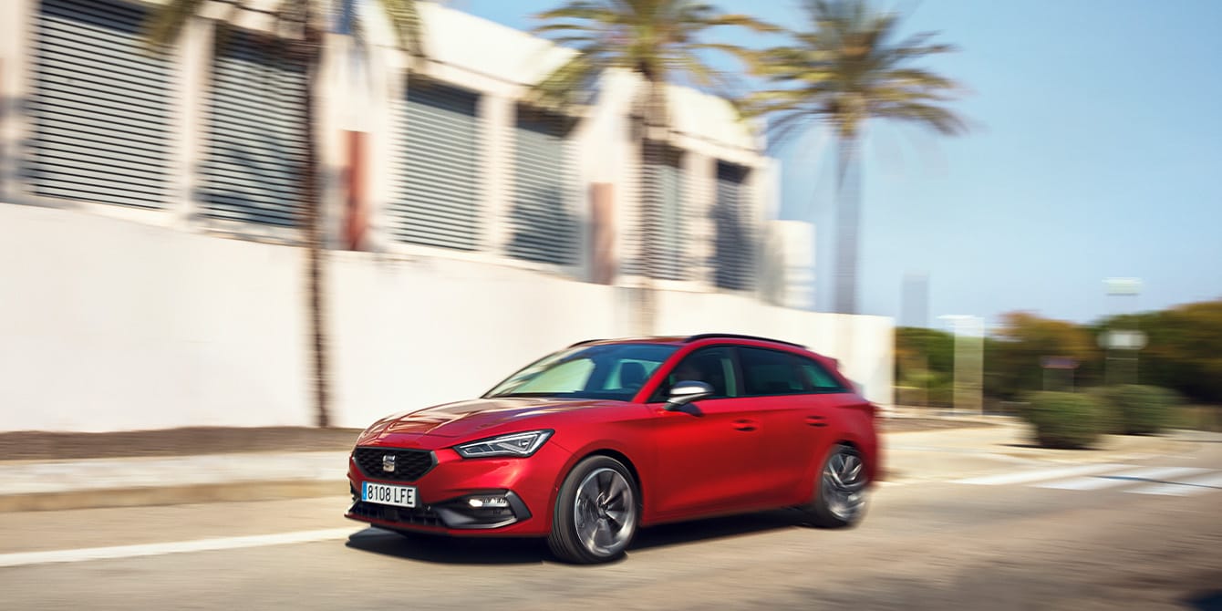 Czerwony SEAT Leon Sportstourer w ruchu, mijający domy i palmy w spokojnej dzielnicy mieszkalnej.