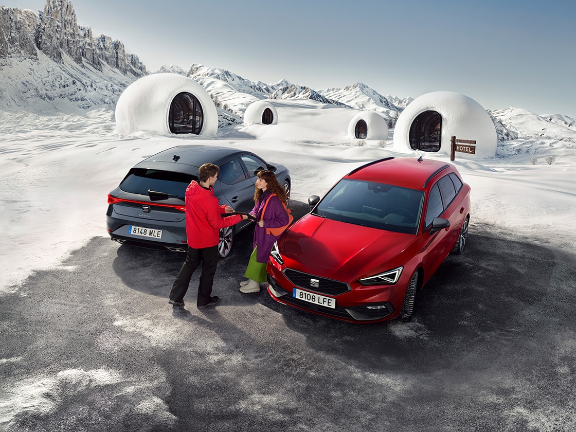 Dwa modele SEAT Leon (5D i Sportstourer) w zaśnieżonej górskiej scenerii, z dwiema osobami pomiędzy pojazdami.
