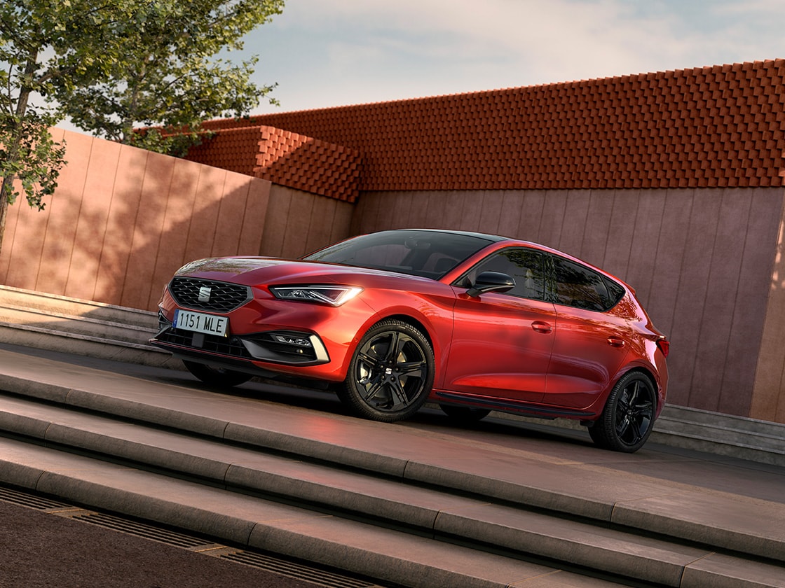 SEAT Leon 5D zaparkowany na zewnątrz, ujęcie z przodu, z budynkiem i drzewem w tle.