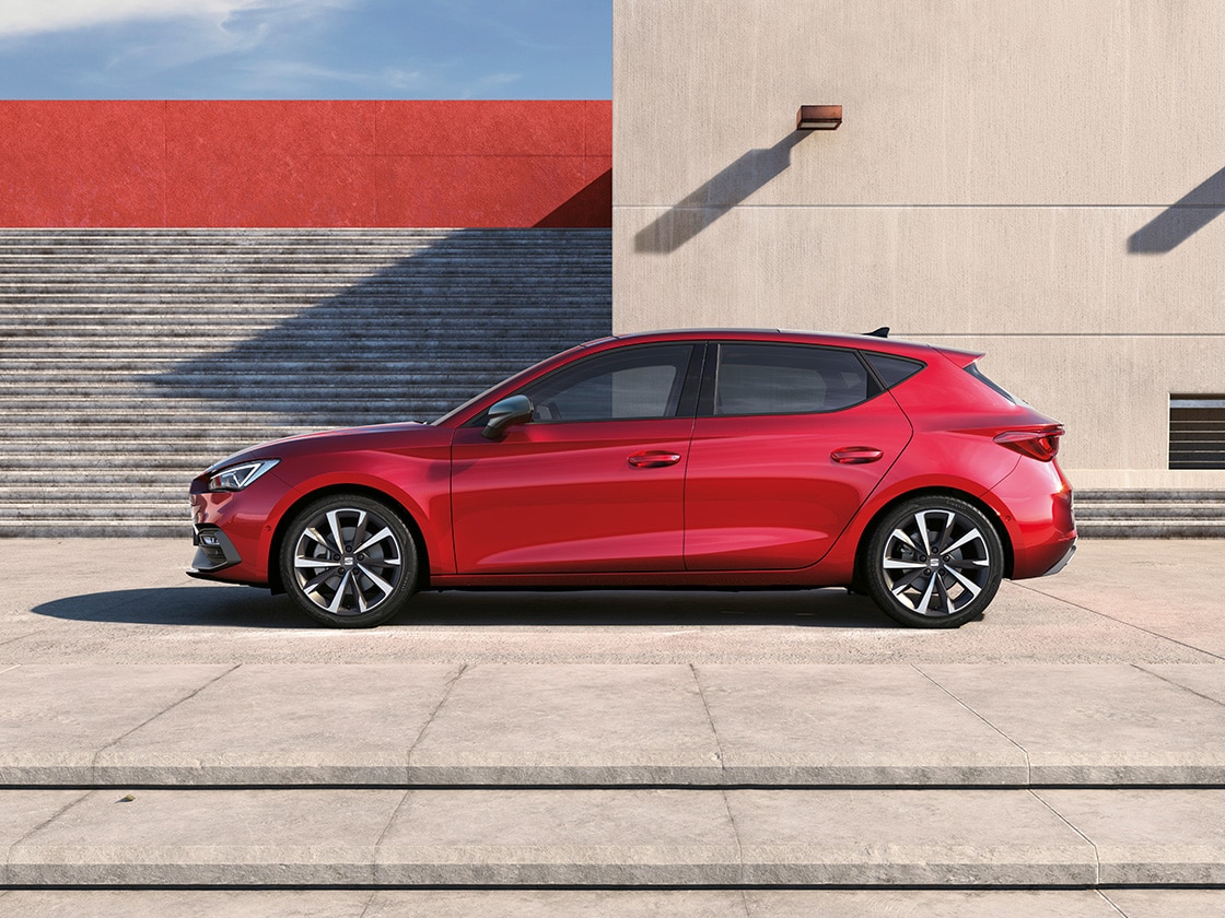 SEAT Leon 5D w kolorze Desire Red, zaparkowany na zewnątrz w słonecznej scenerii, z budynkiem i schodami w tle.
