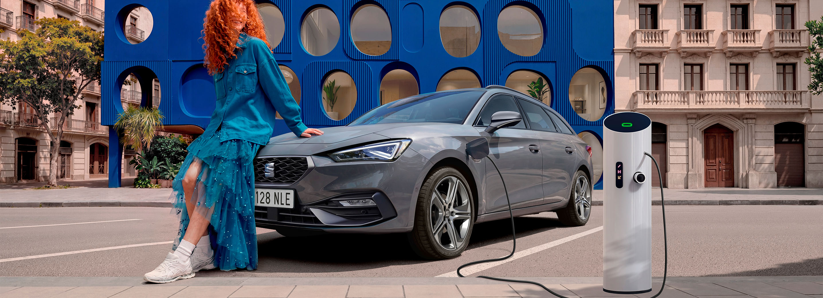 Ujęcie SEAT-a Leon Sportstourer z przodu pod kątem 3/4, w plenerowej, miejskiej scenerii, z budynkami i niebieską konstrukcją w tle.