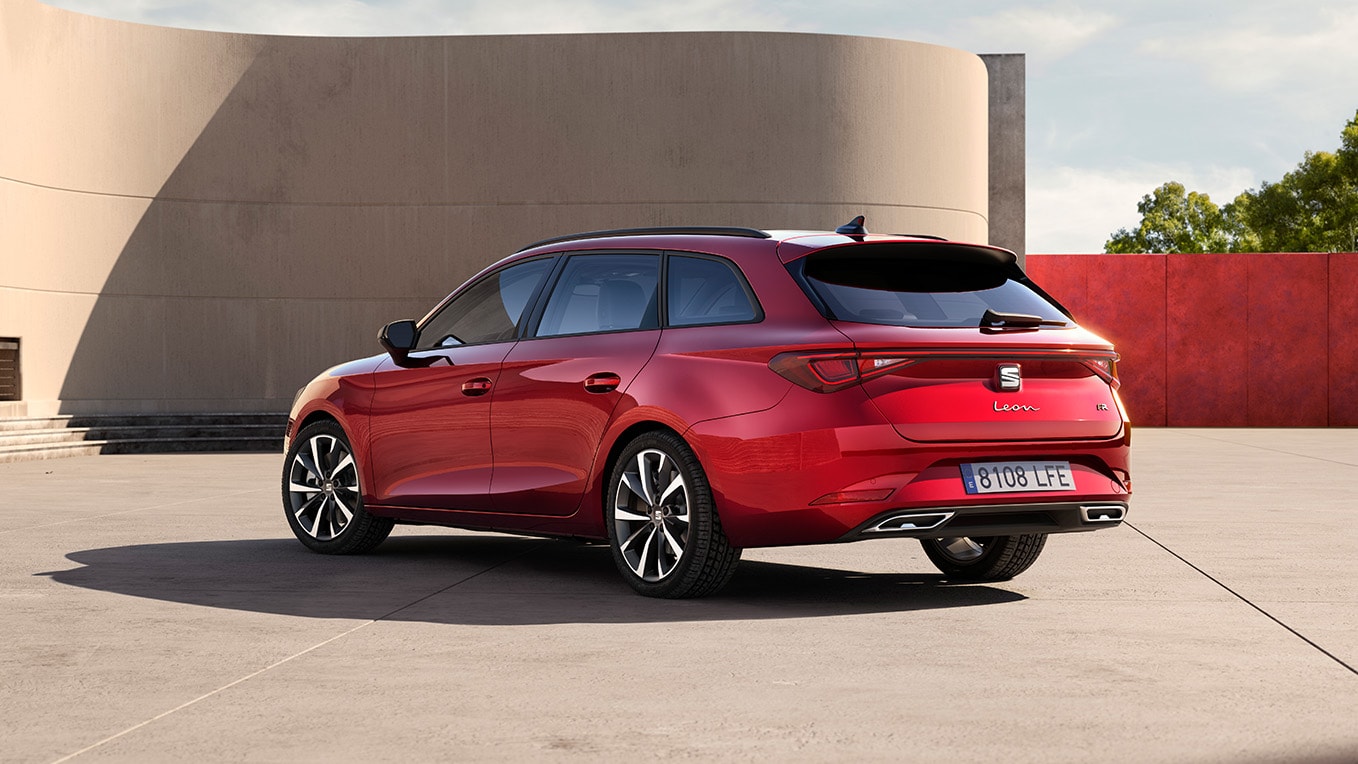 Boczny widok z tyłu samochodu kombi seat leon sportstourer w kolorze desire red, zaparkowanego przodem do ściany budynku. Widoczna tylna część samochodu.