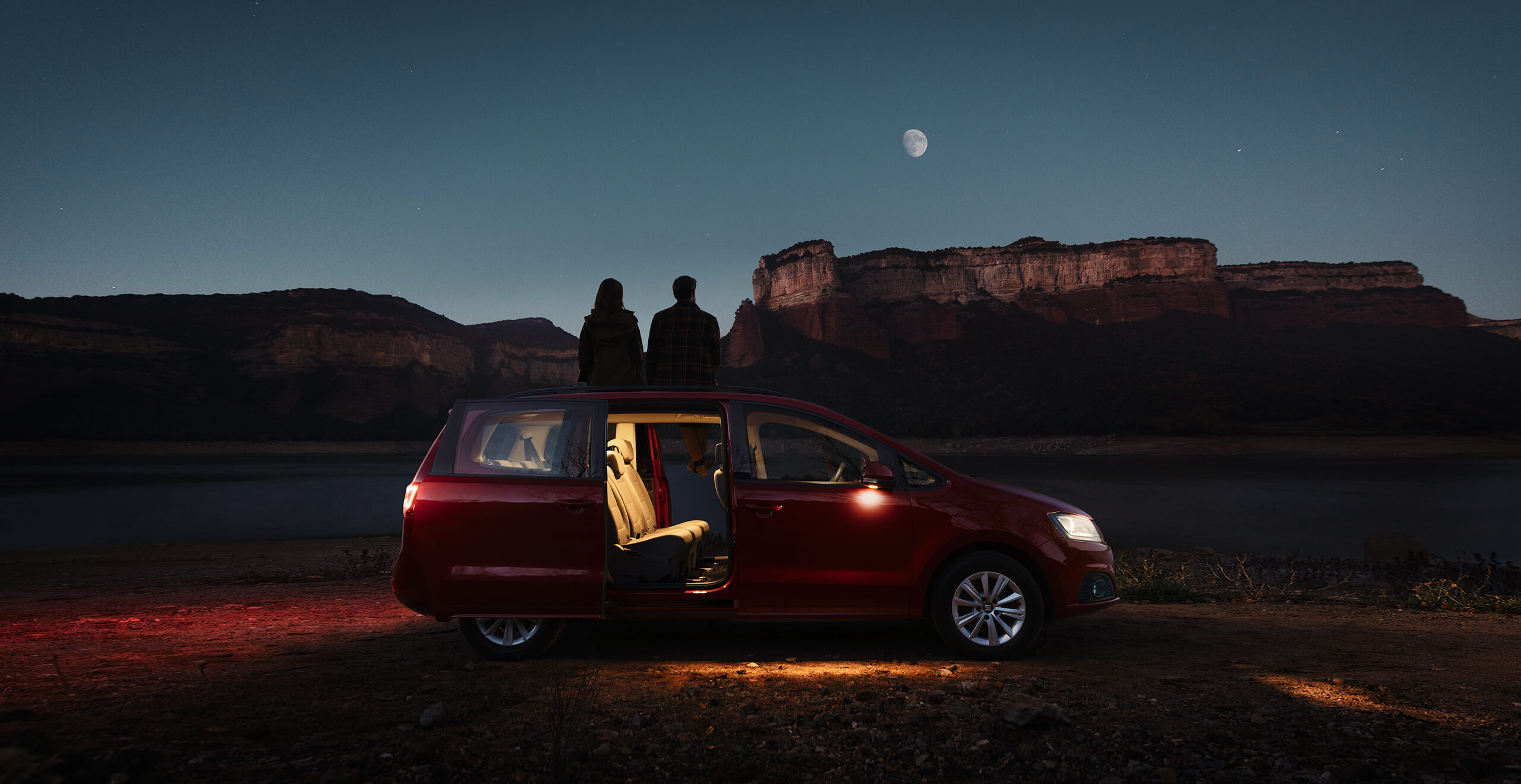 Dwójka ludzi oglądająca księżyc i gory nocą siedząc w otwartych drzwiach SEAT-a Alhambra