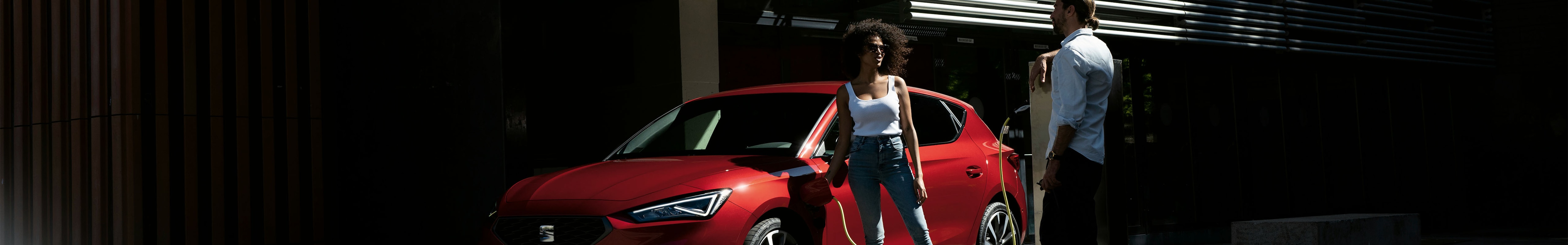 seat leon w kolorze czerwonym i para dyskutująca o tym, czy warto kupić samochód hybrydowy, niski kąt i widok z przodu w trzech czwartych.